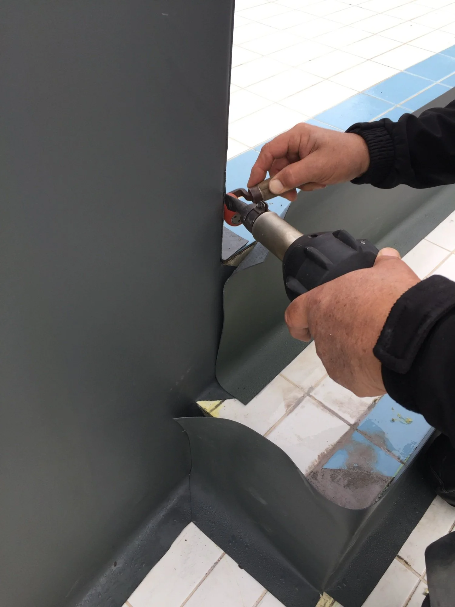 Une personne utilisant une perceuse pour fixer ou assembler un panneau métallique sur un terrain couvert de carreaux blancs et bleus.