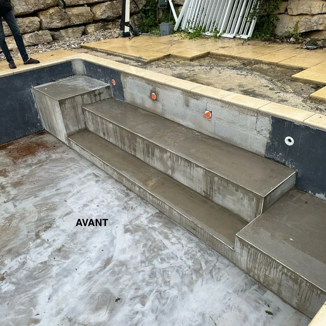 Une piscine en cours de construction avec des escaliers en béton, vue d'un côté. La zone autour est en travaux avec des surfaces en béton et des plantages. Le mot 'AVANT' est écrit sur le sol.
