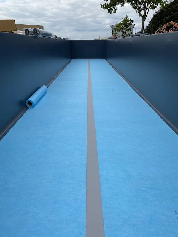 Une piscine vide en cours d'installation avec une nappe bleue et des rouleaux de liner bleus autour.