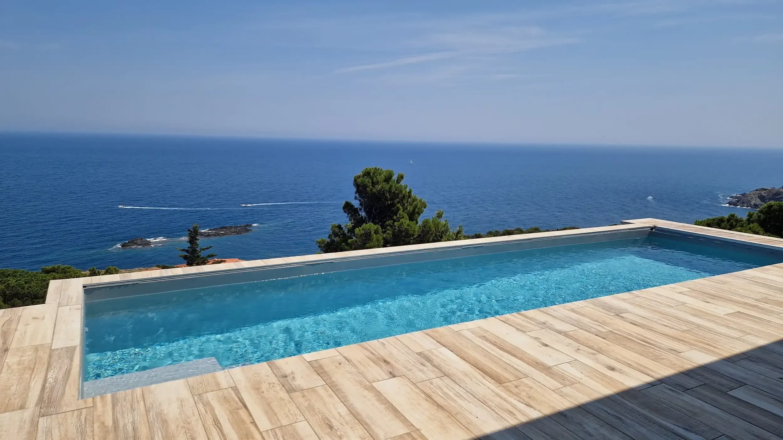 Pose de Pvc armé pour piscine à débordement avec vue sur la mer et des rochers, ciel clair, mobilier de terrasse en bois clair.