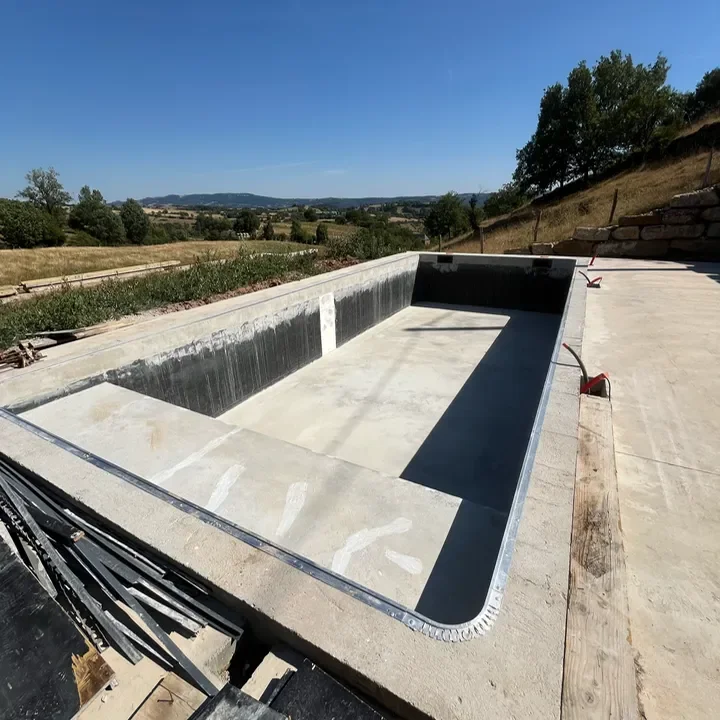 Une bassin rectangulaire vide en cours de construction — avec pose de PVC armé — entourée de campagne et d'arbres par une journée ensoleillée.