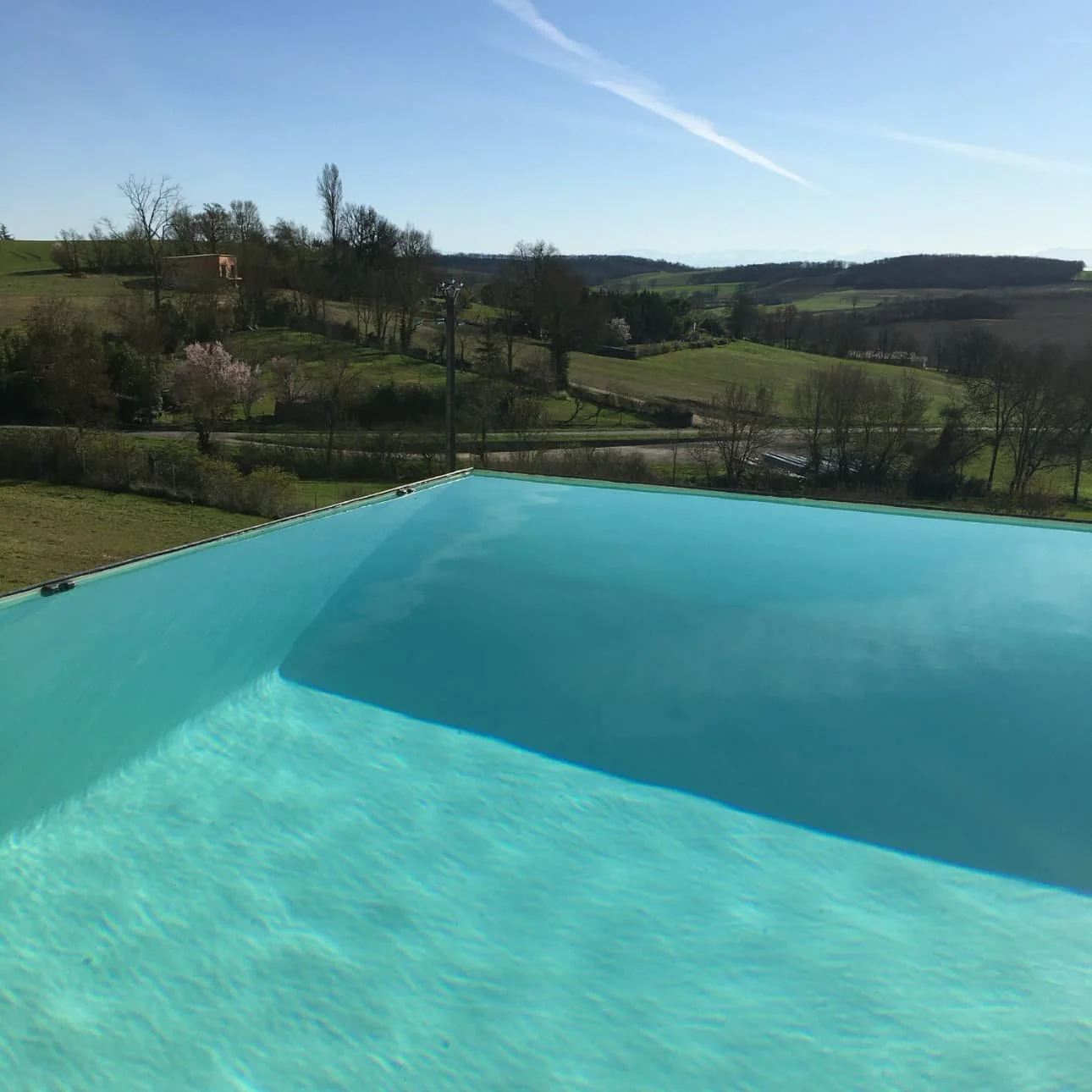 Piscine à débordement avec vue sur un paysage rural verdoyant, ciels bleu clair, arbres et collines en arrière-plan.