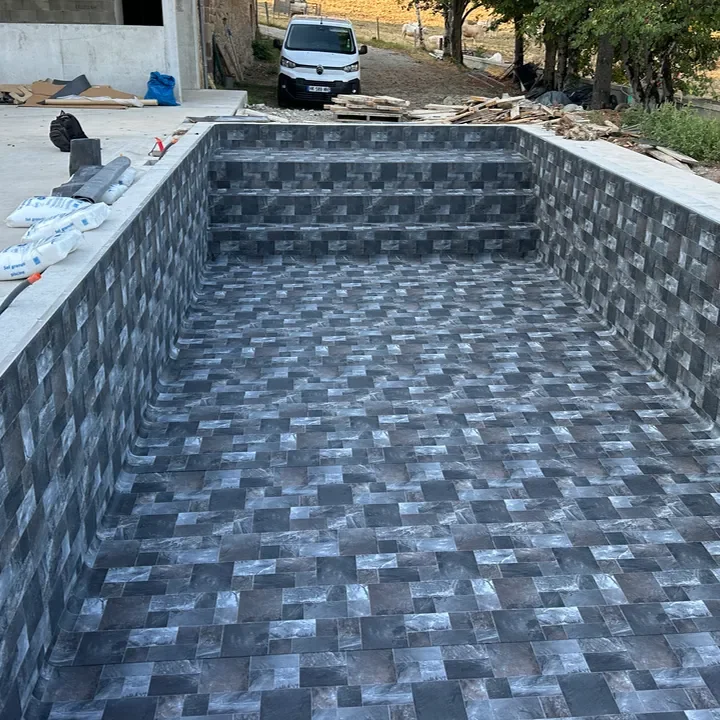 Une piscine en construction avec des murs et un sol carrelés dans des tons gris et noirs, sur un site en extérieur avec des matériaux de construction autour.