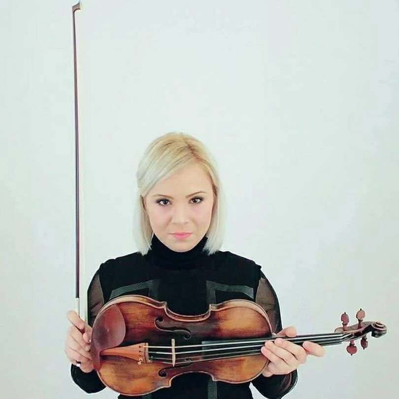 A woman with blonde hair holding a violin and a bow, looking directly at the camera against a plain light-colored background.