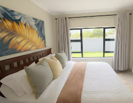 A neatly made bed with white bedding and beige and white pillows in a bright bedroom with large windows and light-colored curtains.