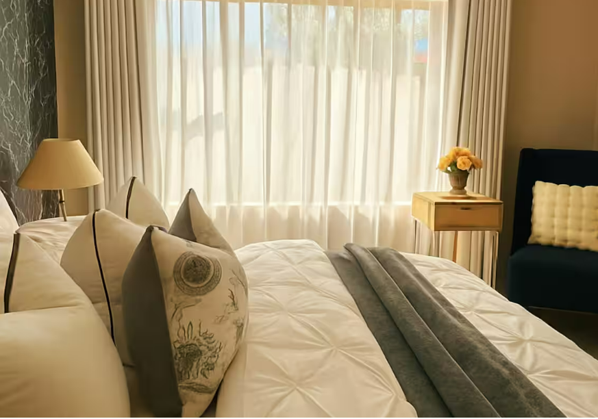 A neatly made bed with decorative pillows, a beige bedside table with a vase of yellow flowers, a black upholstered chair with a white pillow, and a window with sheer curtains in a well-lit bedroom.