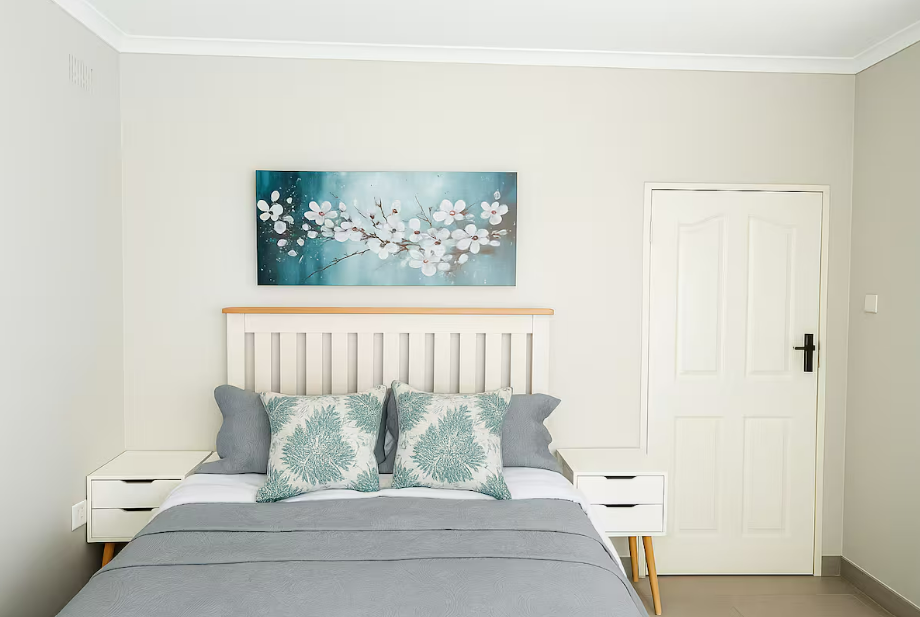 A neatly made bed with white and light teal bedding, two large decorative pillows with botanical patterns, two smaller gray pillows, a white nightstand on each side, a painting of white blossoms on a blue background above the bed, a white door with a