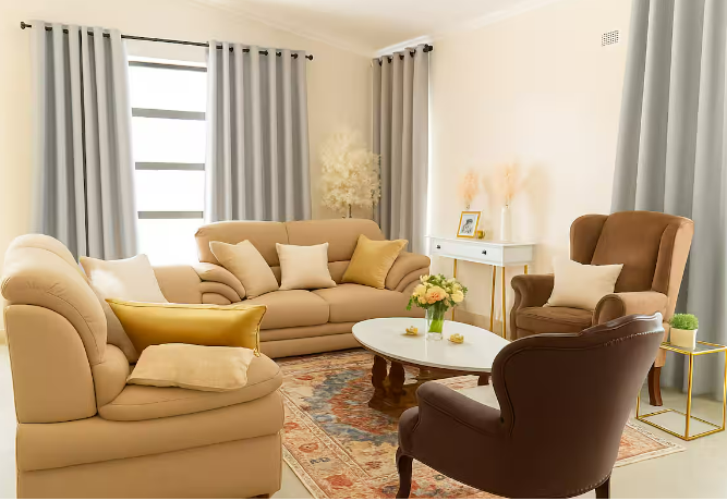 Living room with beige and brown furniture, including a sofa, armchair, and a tufted armchair, with pillows. There is a white coffee table with a flower arrangement, a side table, and a white console table against the wall. The room has light-colored