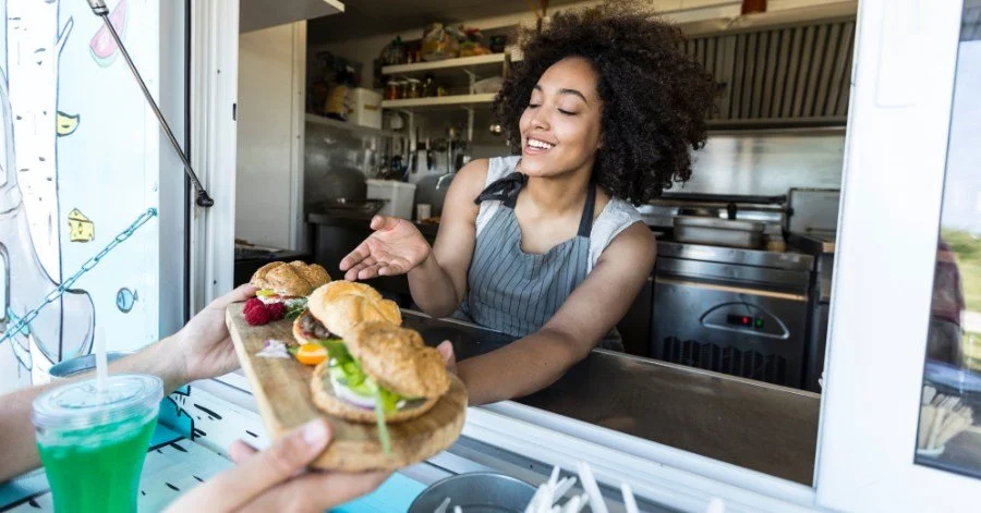 A woman in a food truck is happily accepting a wooden platter with a sandwich, raspberries, and a small drink from a customer.