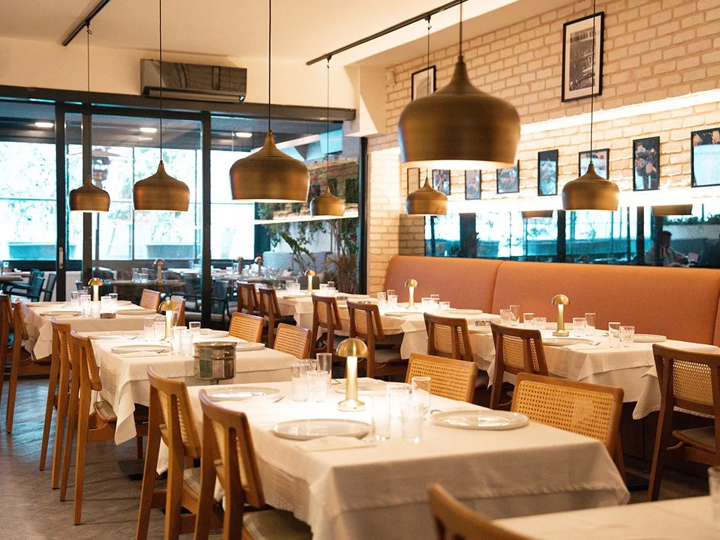 A stylish restaurant interior with dining tables covered in white tablecloths, set with plates, glasses, and small lamps, featuring warm lighting, a brick wall with framed photos, and large windows.