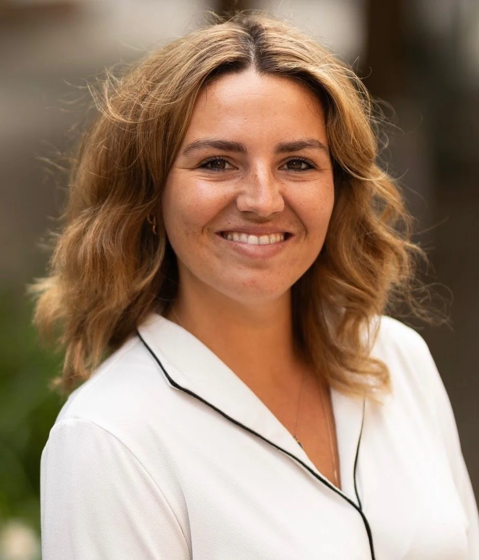 Portrait of a Tansy Parkinson who is a psychodynamic therapist with light brown, curly hair wearing a white shirt with black piping, standing outdoors.