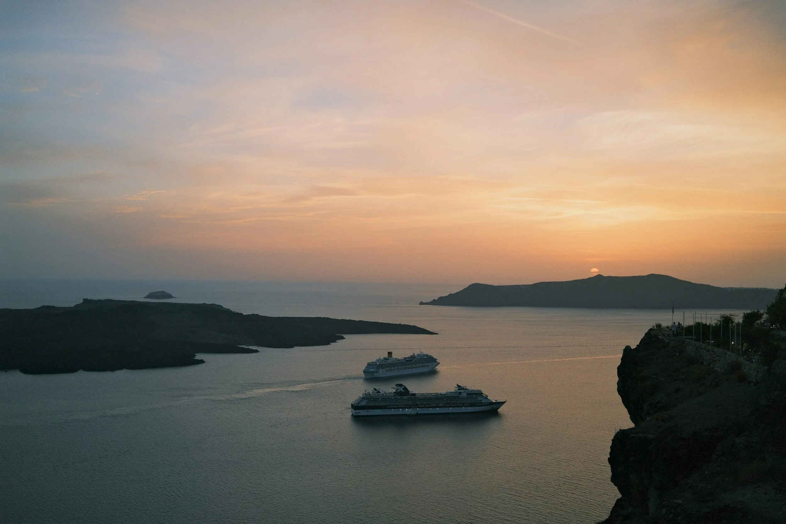 Sunset over a harbor with two cruise ships, rocky cliffs on the right, and distant islands in the background.
