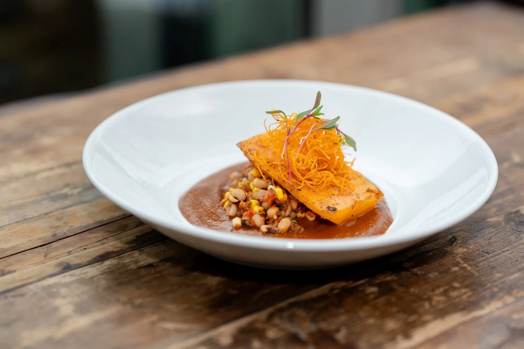 A plated dish featuring grilled squash topped with shredded carrots and microgreens, served with a side of mixed beans in a sauce.