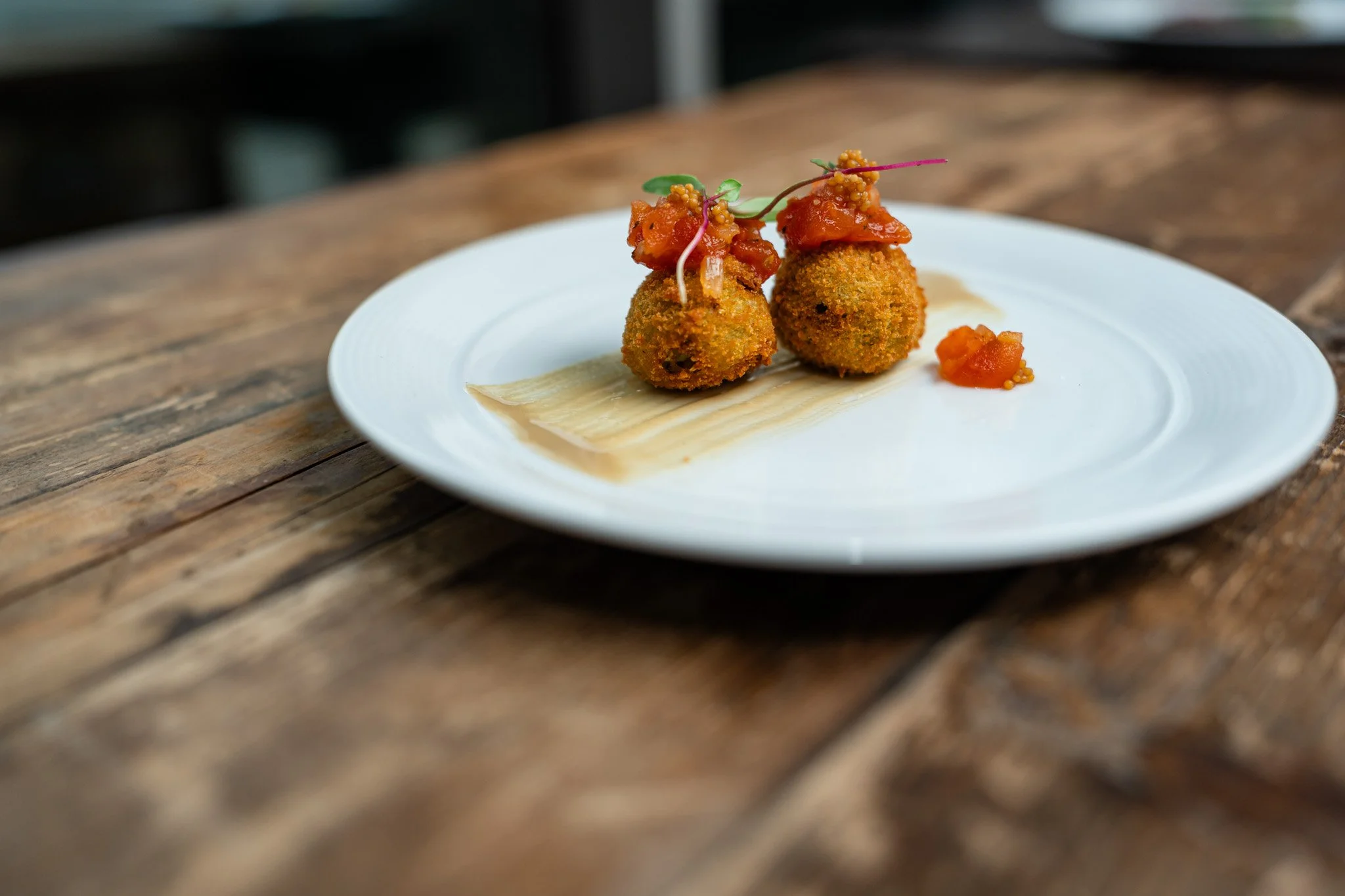 Fried croquettes topped with tomato sauce and garnished with microgreens on a white plate, served on a rustic wooden table.