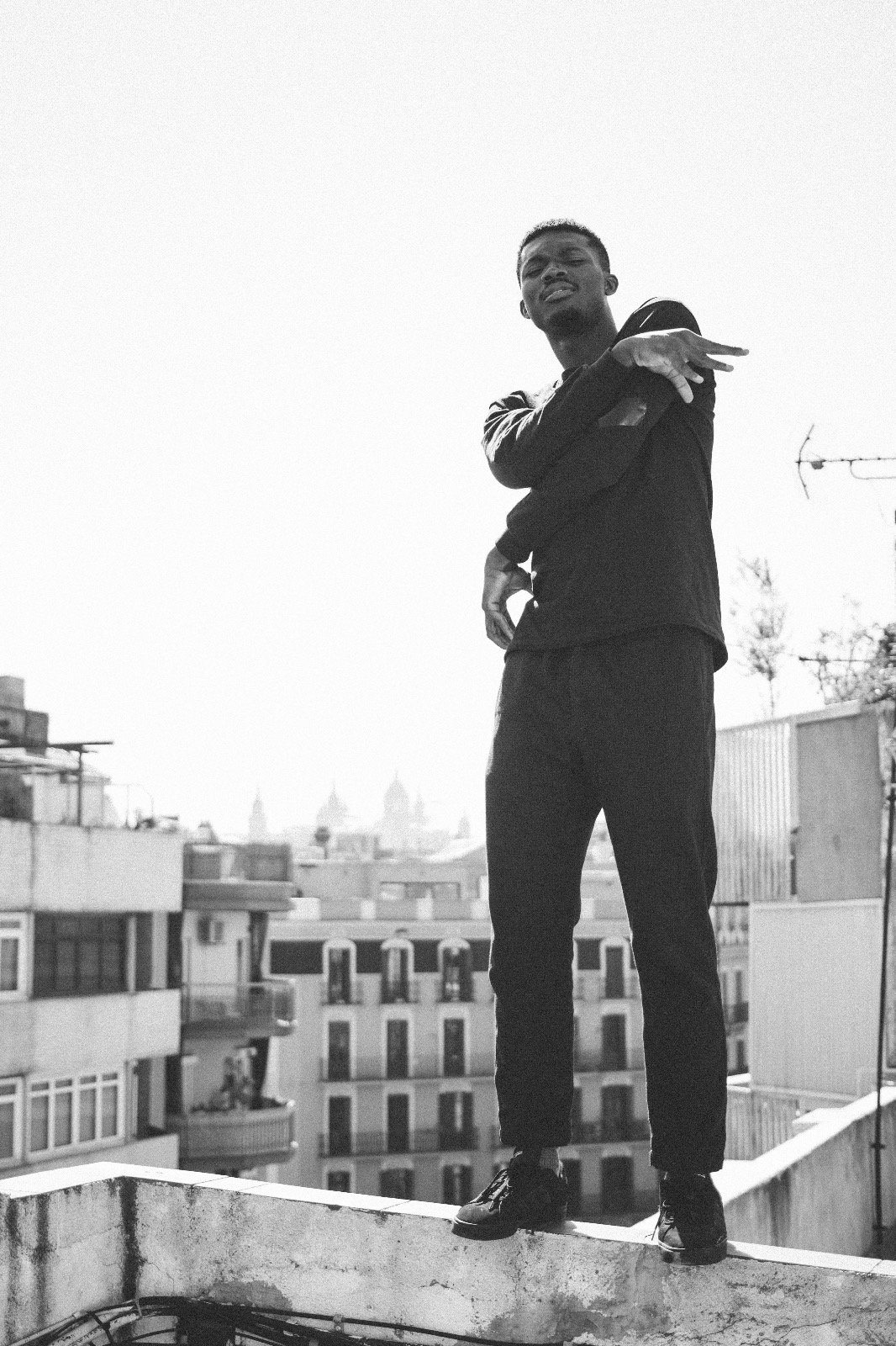 A young man standing on a rooftop in an urban area, making a gesture with his right hand, wearing a black jacket and pants, with city buildings in the background.