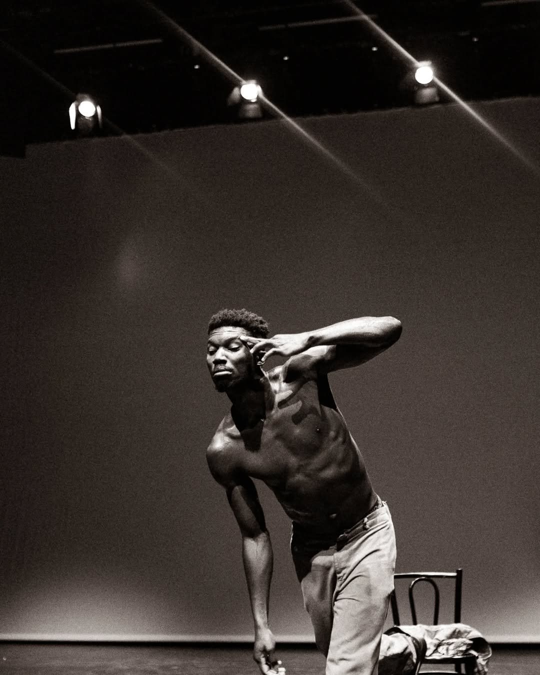 A shirtless man performing on stage, leaning forward with one hand near his face and the other holding an object, with a chair in the background.