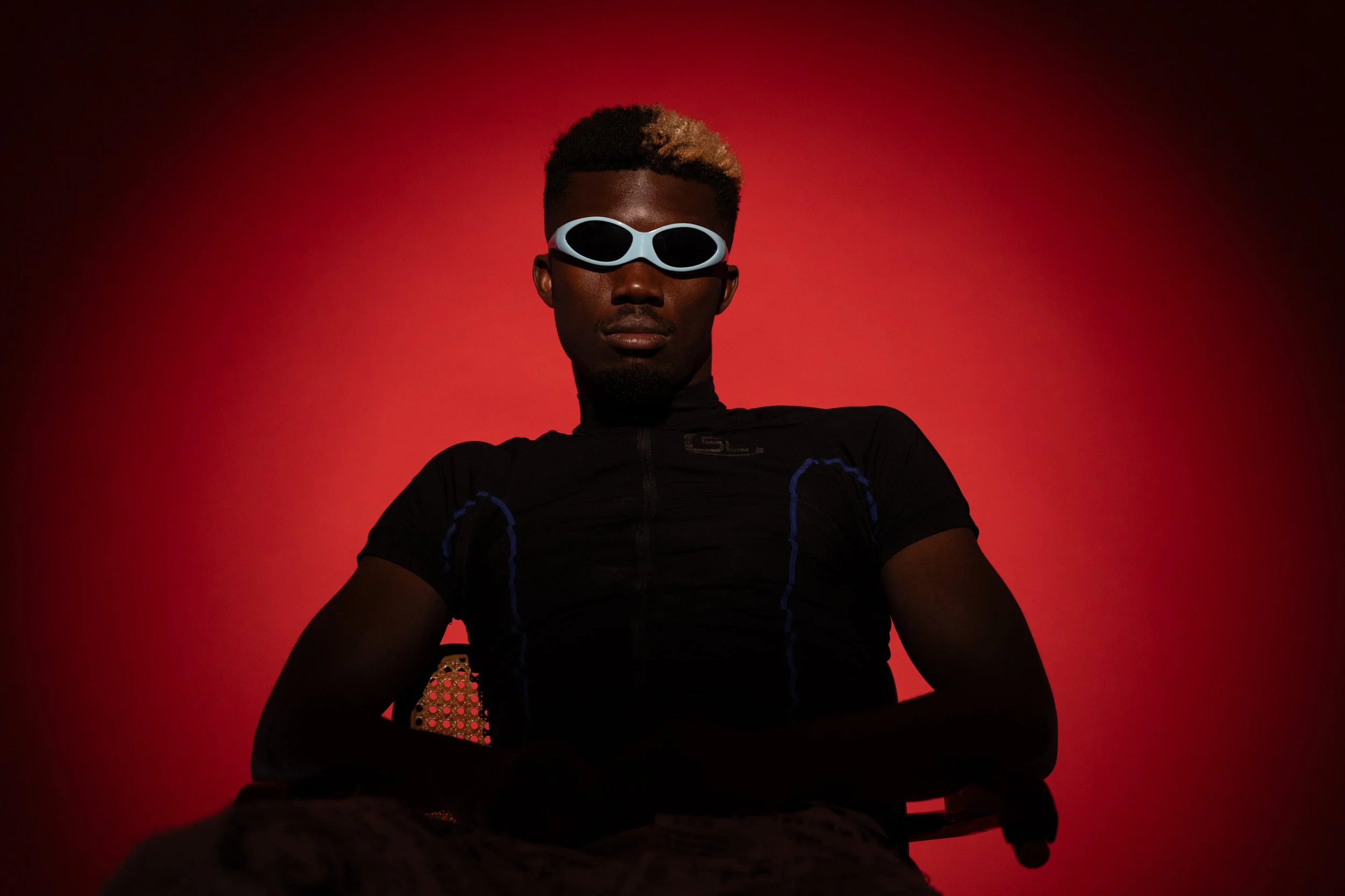 Teenage boy wearing black athletic shirt and futuristic white sunglasses sitting against a red background.