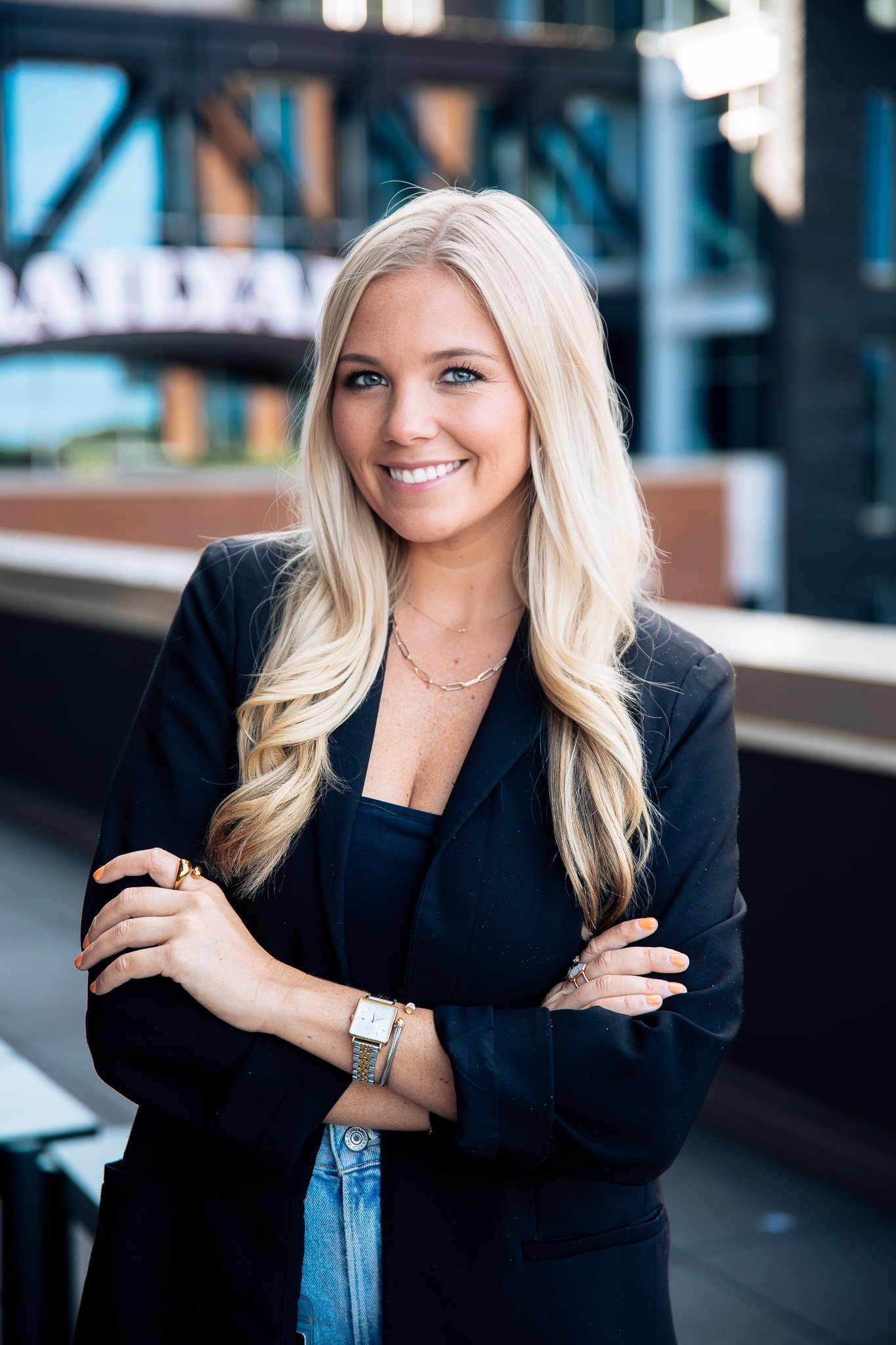 A smiling woman with long blonde hair, wearing a black blazer, jewelry, and a watch, standing outdoors in an urban environment.