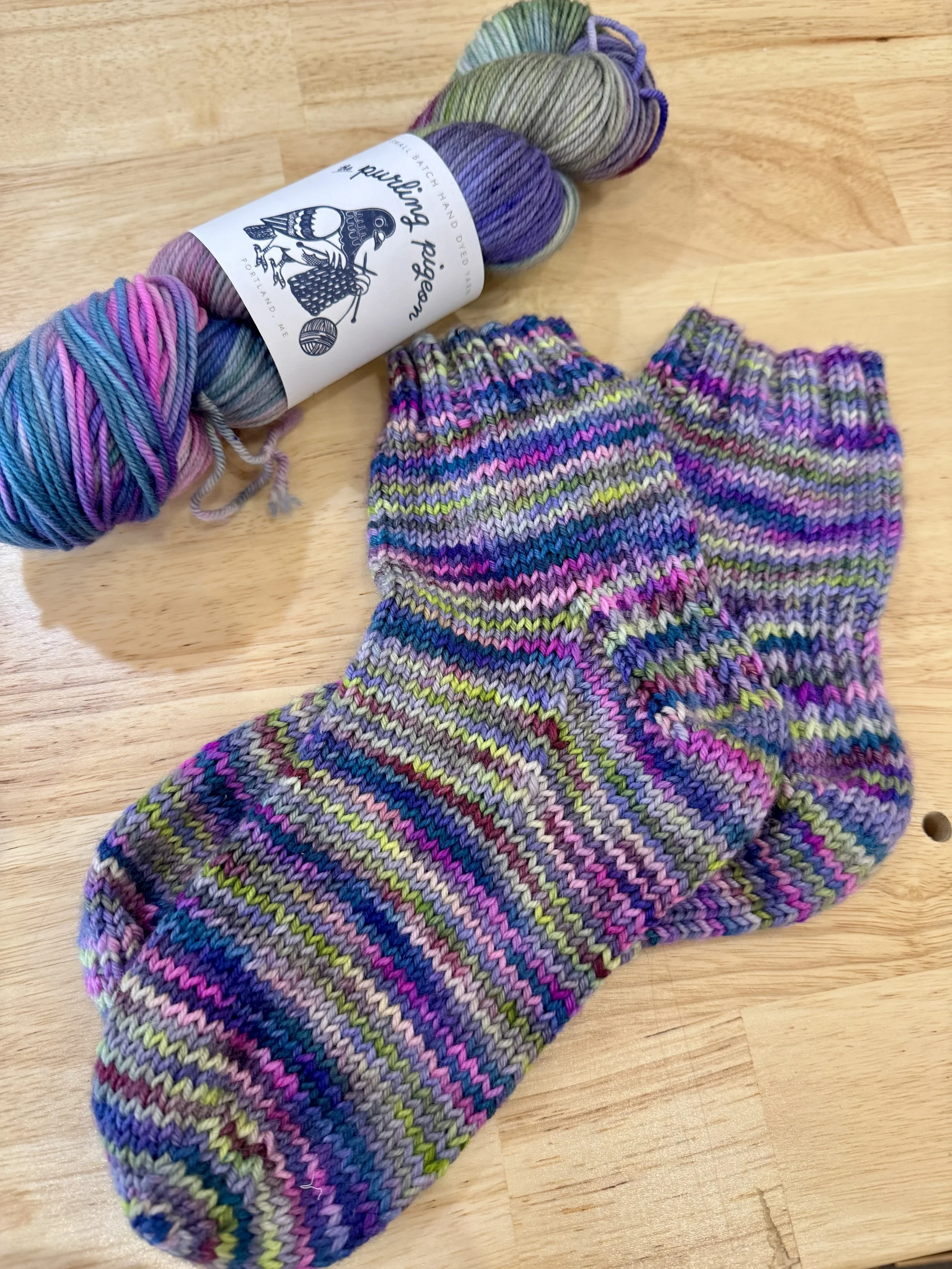 A pair of colorful striped knitted socks placed on a wooden surface with a skein of multicolored yarn nearby.
