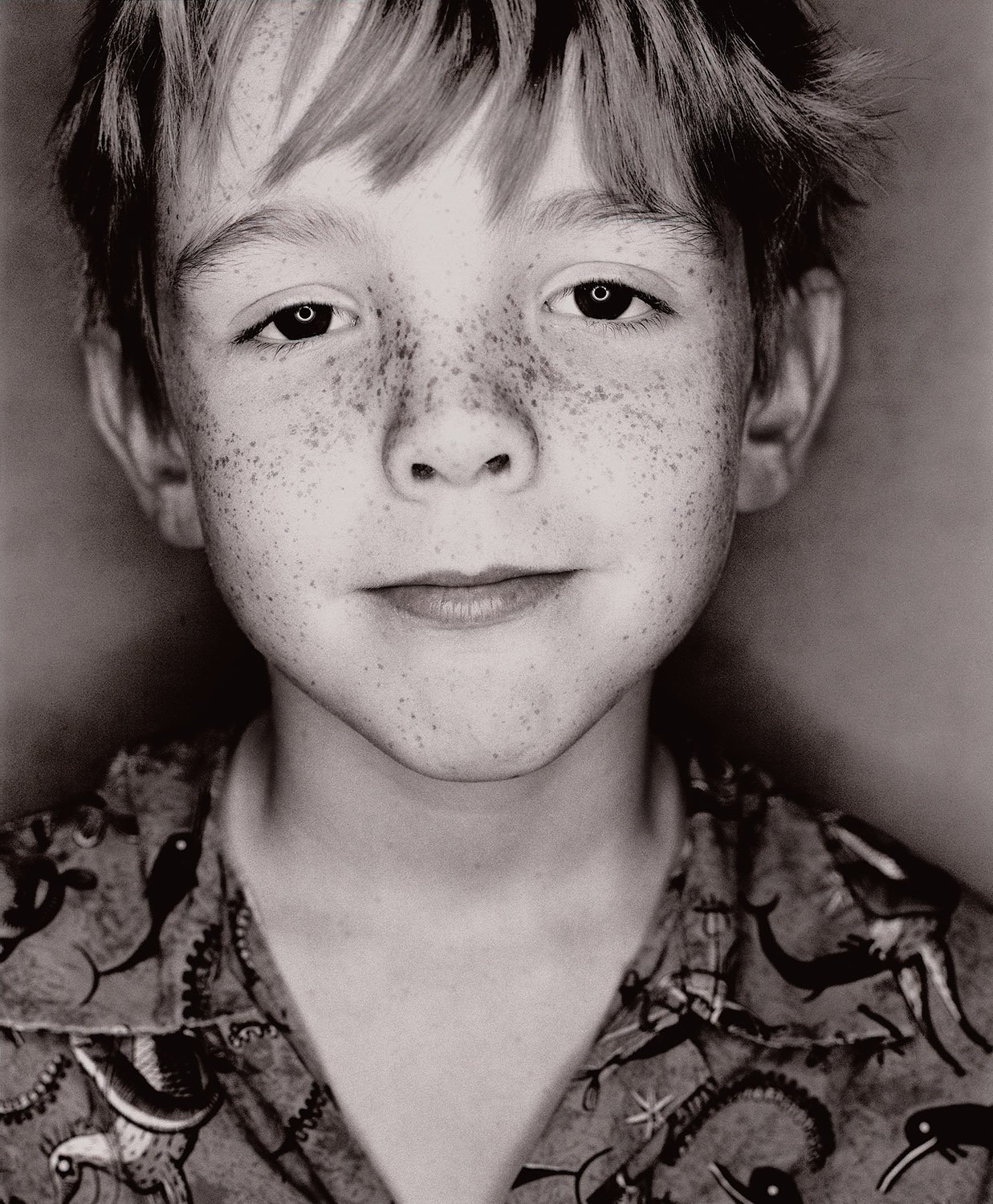 Close-up black and white photo of a young boy with freckles, smiling slightly, wearing a patterned shirt.