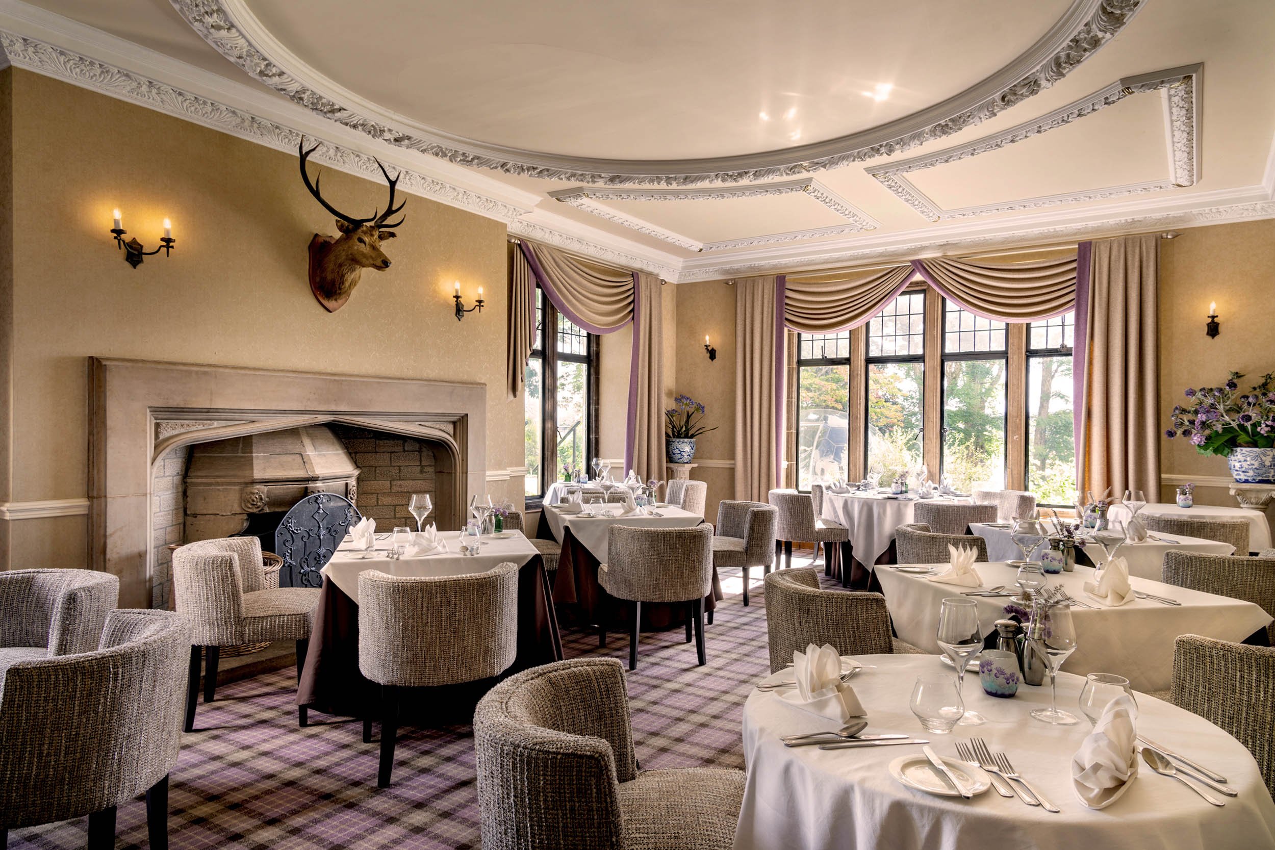 Elegant restaurant interior with large windows, beige walls, and a decorative ceiling. A mounted deer head with antlers is above a fireplace, flanked by wall sconces. The room features tables set with white tablecloths, glassware, and napkins, and is