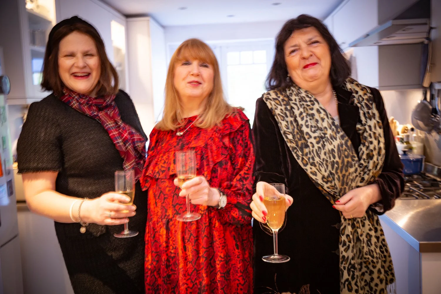 Three women standing in a kitchen, smiling and holding glasses of champagne at a social gathering.