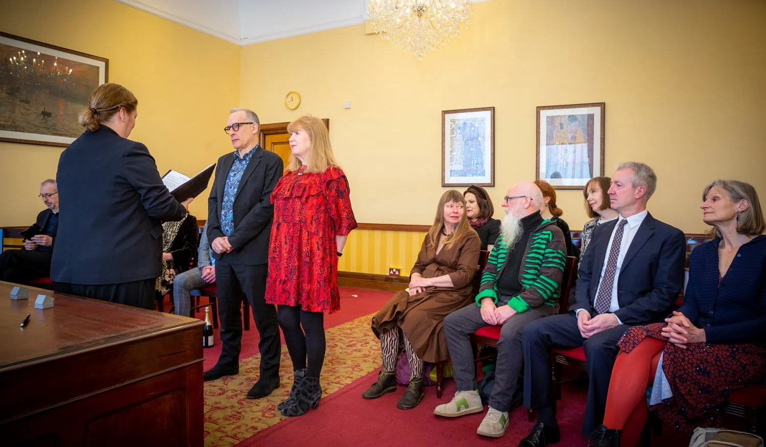 A group of people at a formal event, with three individuals standing and reading from papers or a book and several seated behind them, inside a room with yellow walls, framed artworks, and a chandelier.