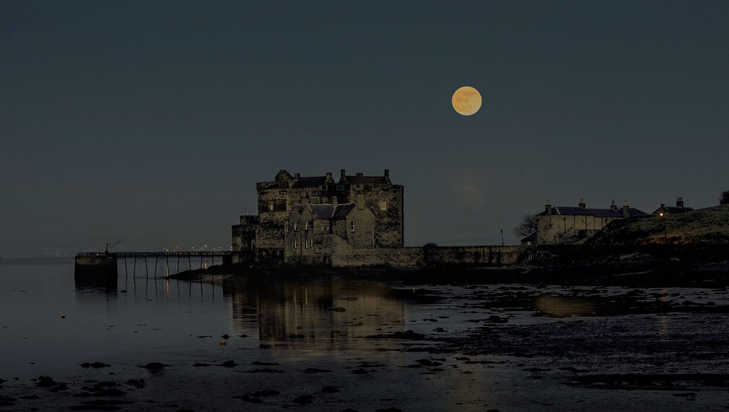 Blackness Castle on the coast at nighttime with a full moon in the sky and reflections on the water.