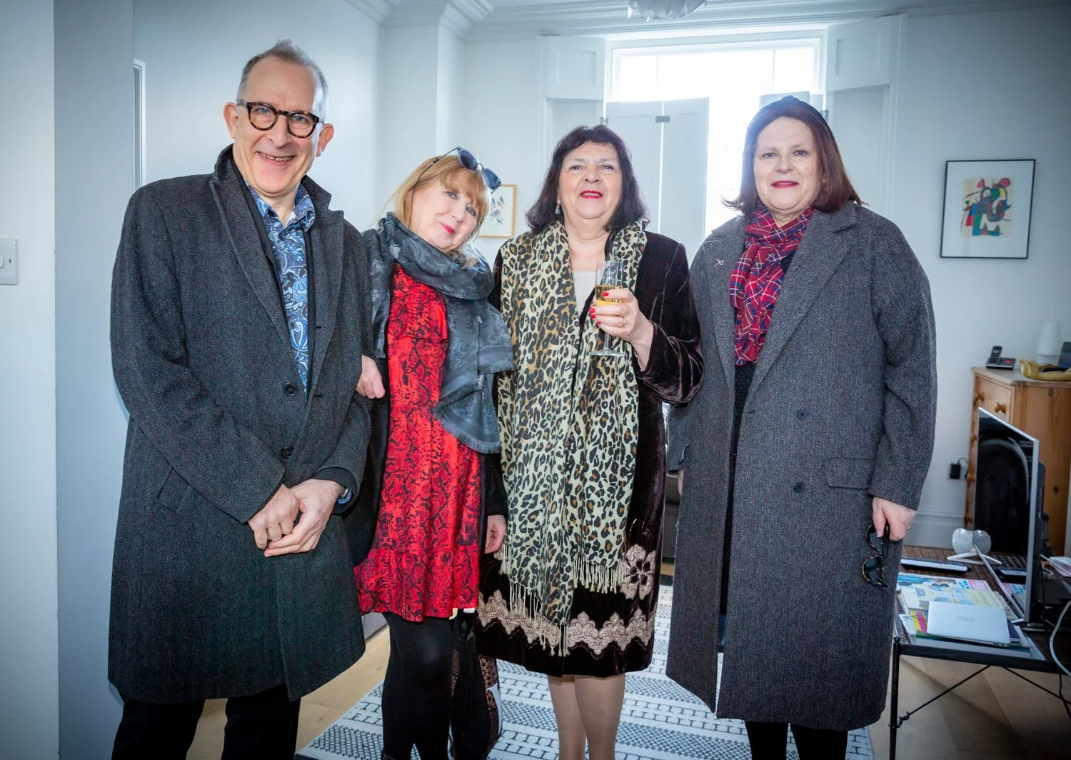 Four people in formal attire standing together in a living room, smiling, with a woman holding a glass of wine.