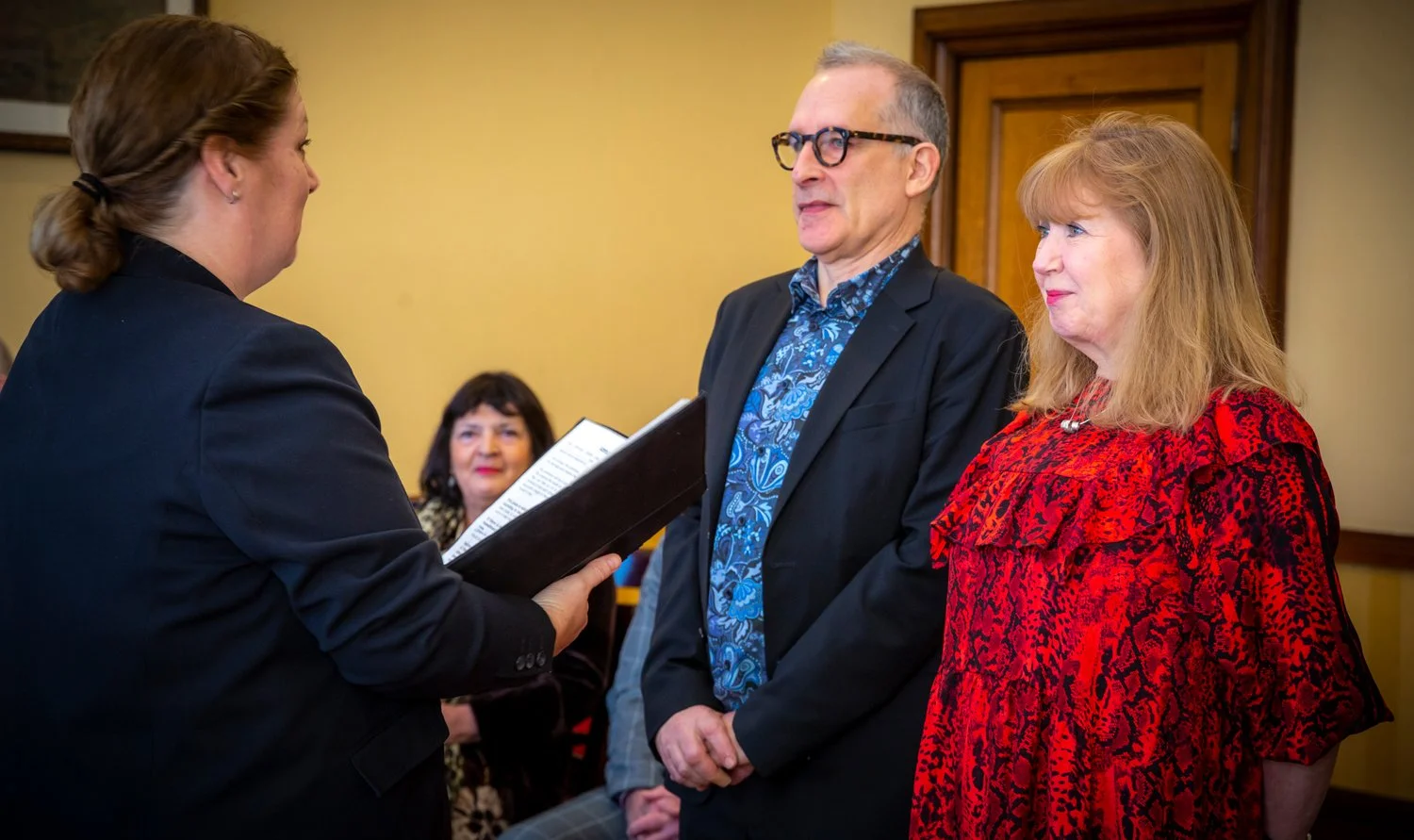 A woman in a black blazer holding a framed certificate presents it to a man and a woman, who are standing side by side; the man wears glasses and a dark blazer with a blue patterned shirt, and the woman has red hair and wears a red patterned dress. I