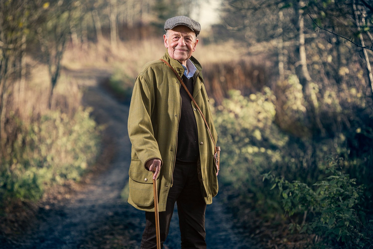 An elderly man walking outdoors on a dirt path in a wooded area, smiling and holding a walking stick. He is wearing a flat cap and a green coat.
