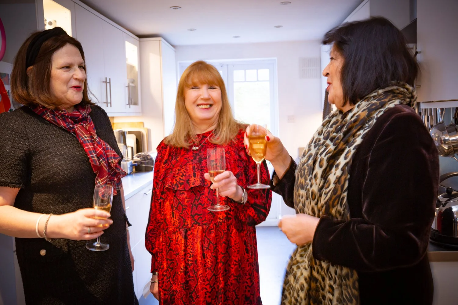 Three women in a kitchen enjoying drinks and conversation, smiling and engaging with each other.