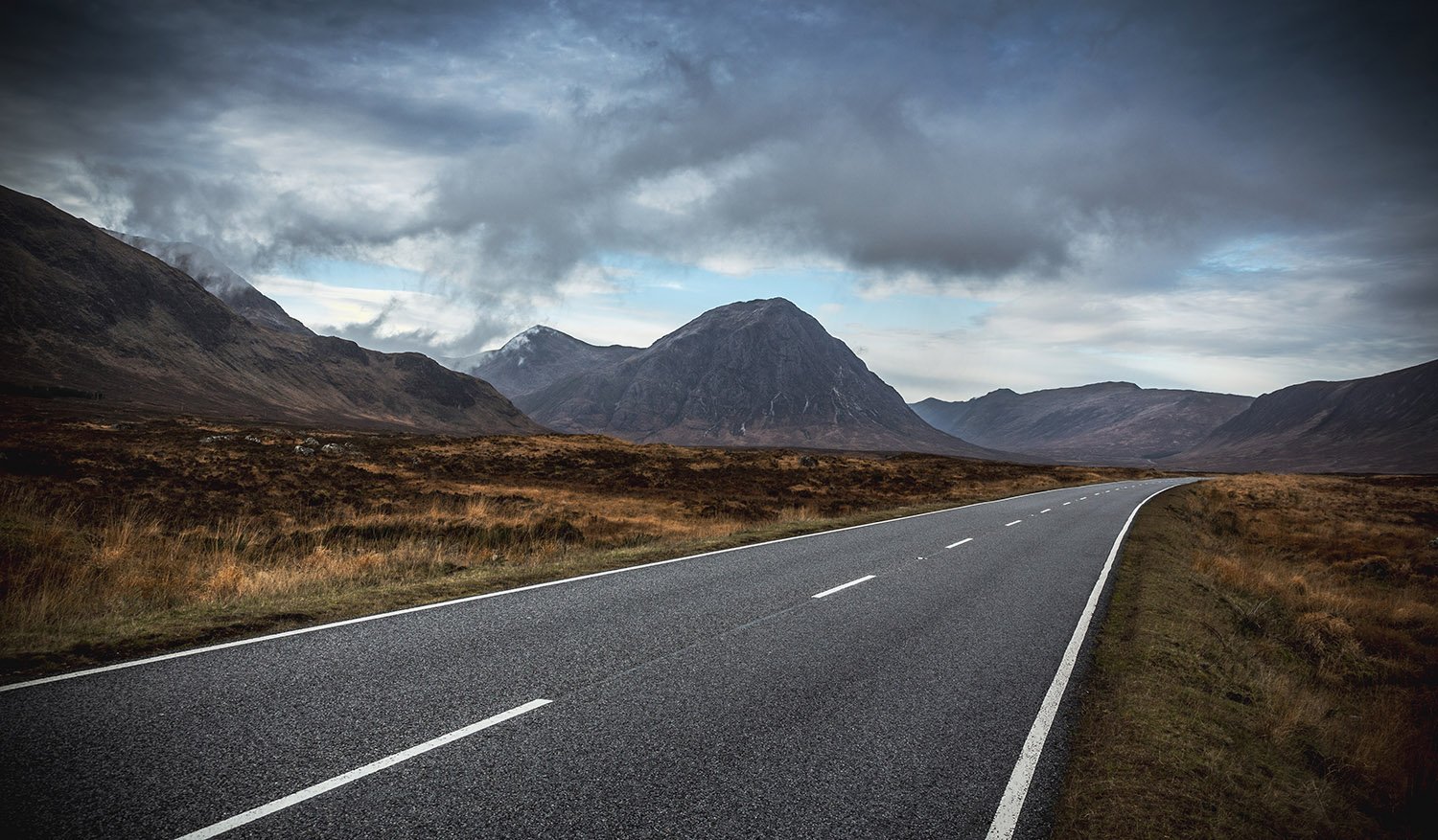 Empty winding Glen Coe road through a mountainous landscape with dark, cloudy sky.