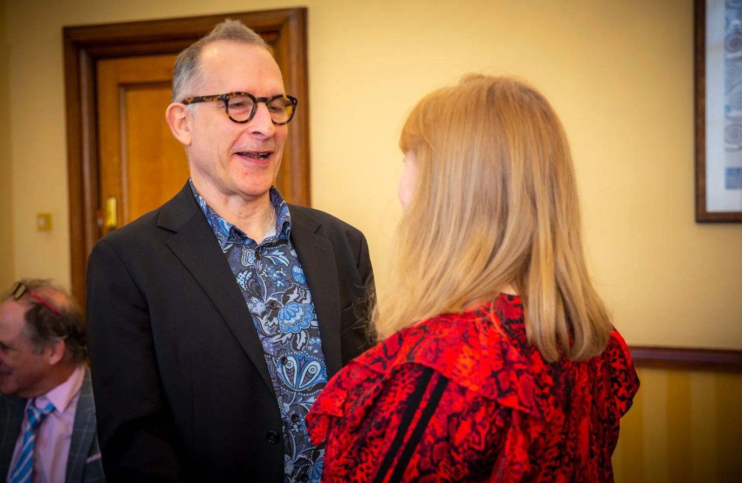 A man with glasses and a dark suit talking to a woman with blonde hair wearing a red and black patterned jacket, in a room with yellow walls and wooden doors.