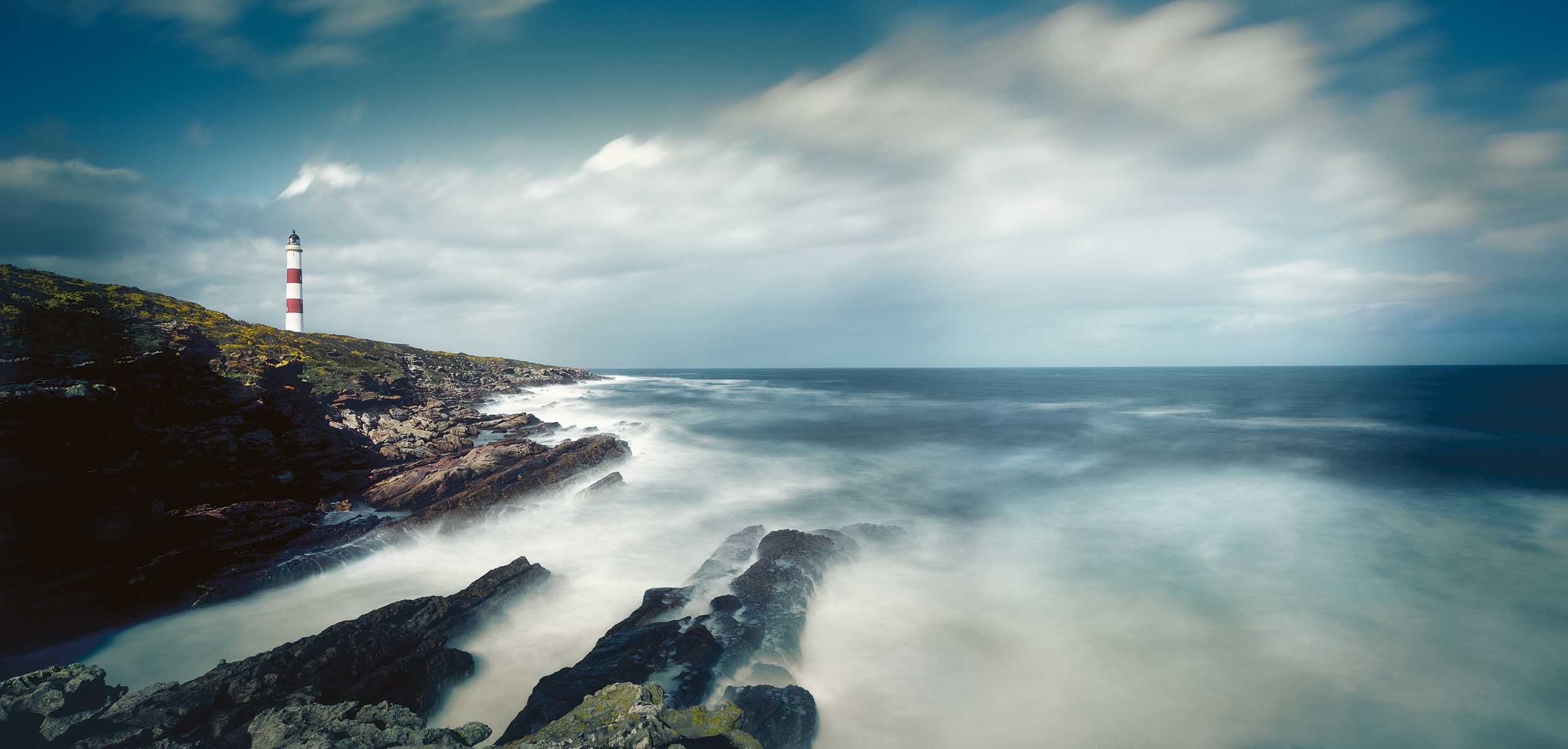 A coastal scene with a lighthouse on a rocky hilltop, ocean waves crashing against dark rocks, and a cloudy sky.