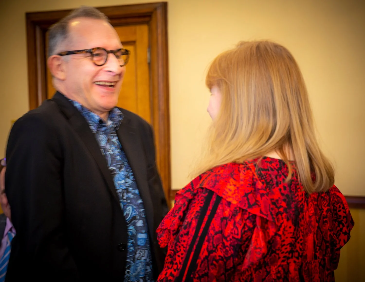 A man with glasses and a woman with red hair laughing and talking indoors.