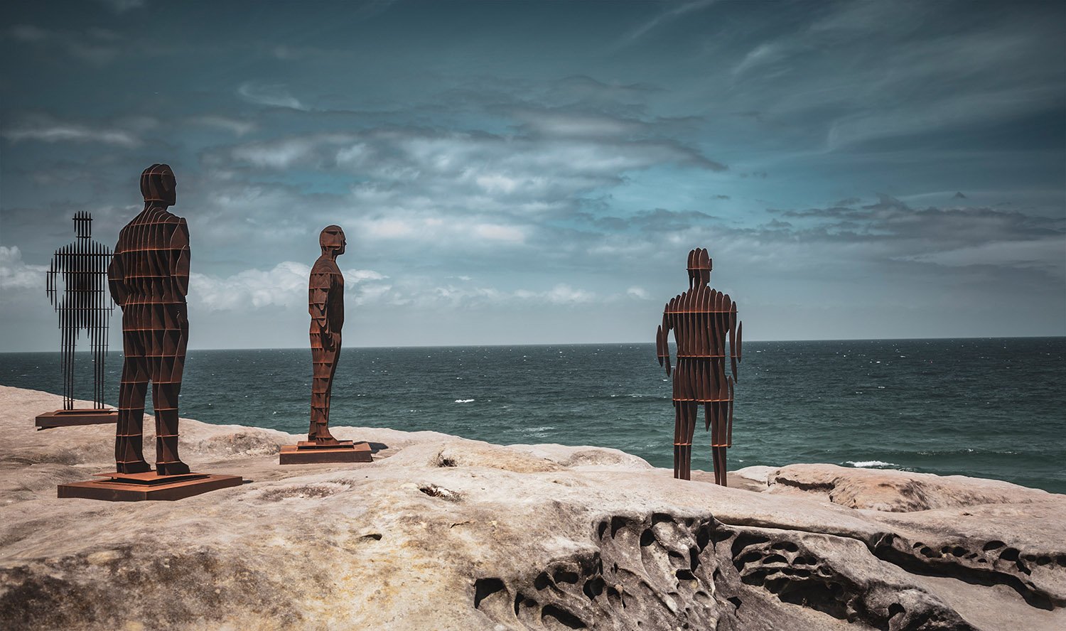 Four metal sculptures of human figures stand on a rocky coastline with the ocean and cloudy sky in the background.