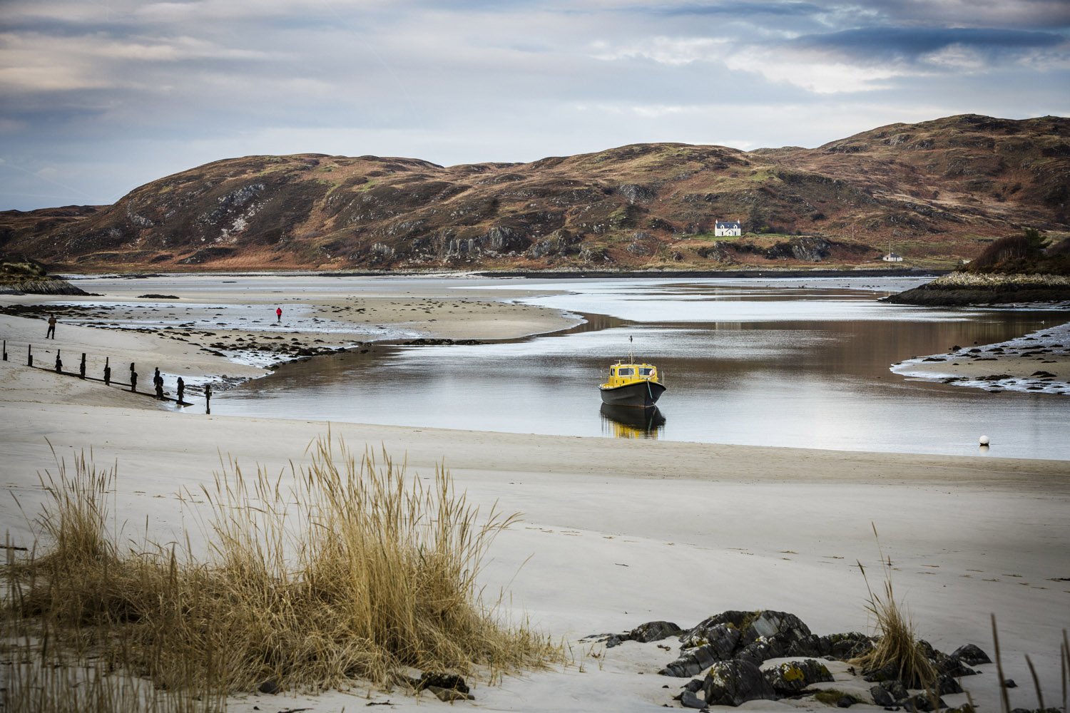 A boat on a calm river or bay, with a sandy beach and grassy dunes in the foreground, and rolling hills with sparse vegetation in the background under a cloudy sky.