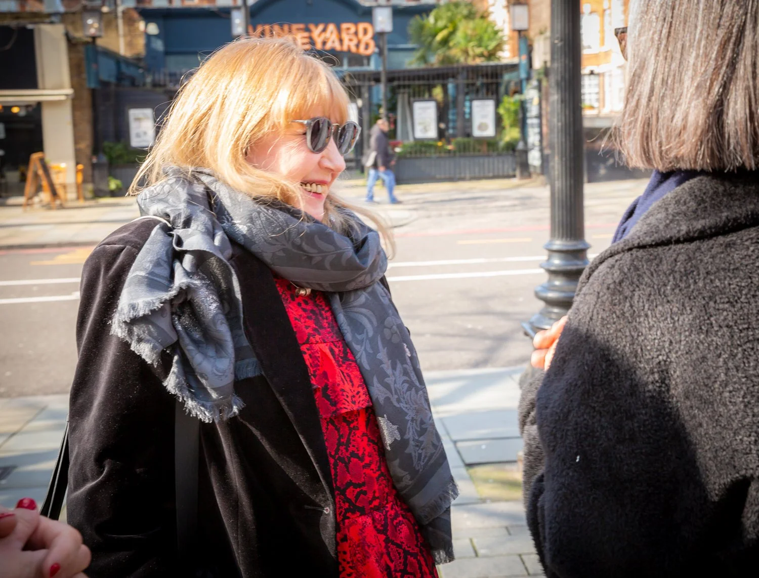 Two women smiling and talking outdoors near a street with pedestrian crossing and streetlamp, with a sign reading 'Vineyard' in the background.