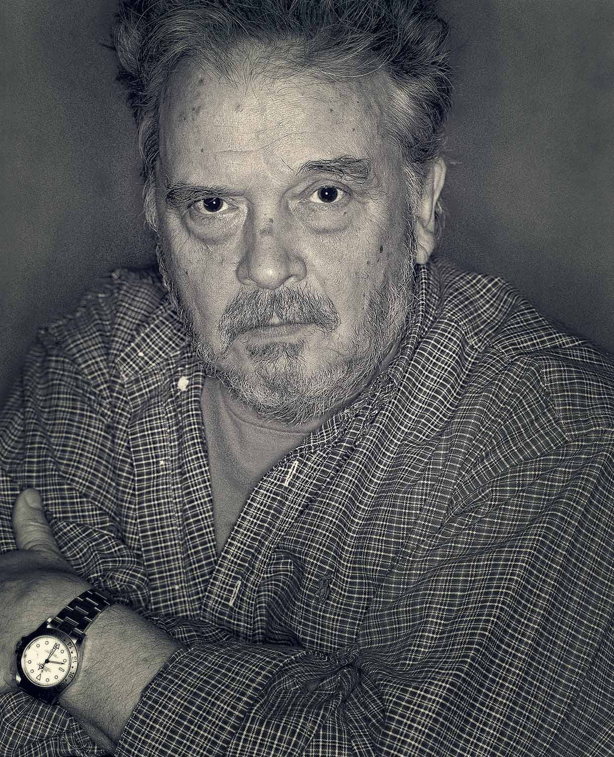 A black and white portrait of Bailey (David Bailey) wearing a checkered shirt and a watch on his left wrist.