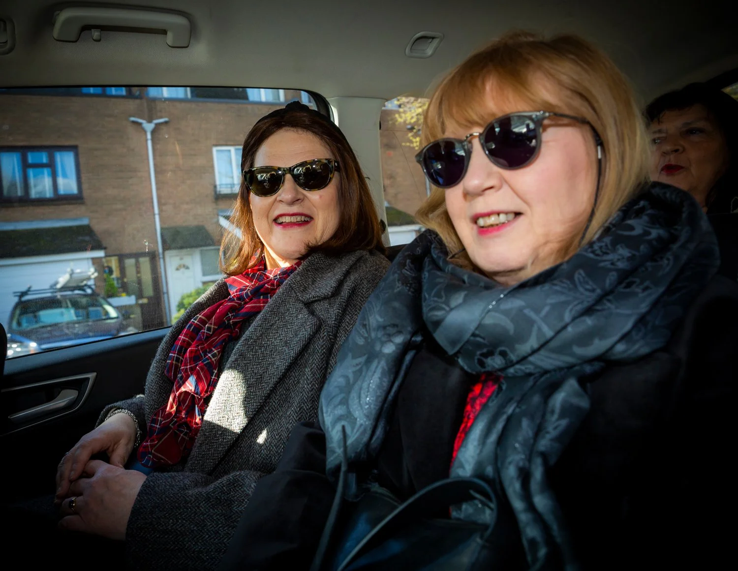 Two women wearing sunglasses sitting in a vehicle, smiling, with a brick building visible outside the window.