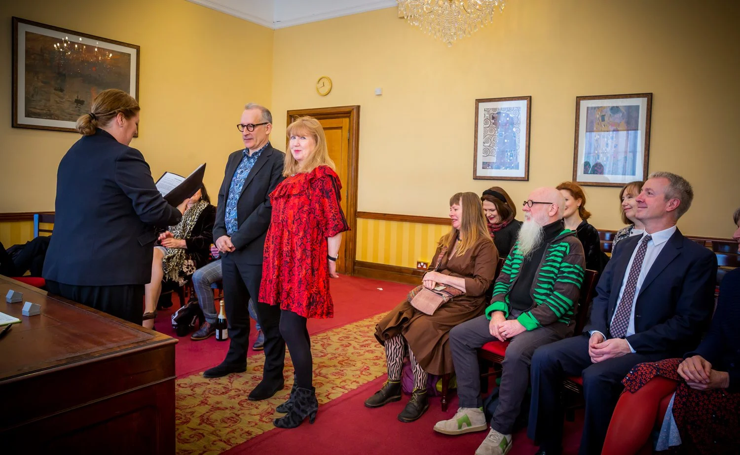 A woman in a black suit is reading a document or certificate during a ceremony, with a woman in a red dress and glasses standing nearby, and a group of seated people watching, in a room with yellow walls and framed art.