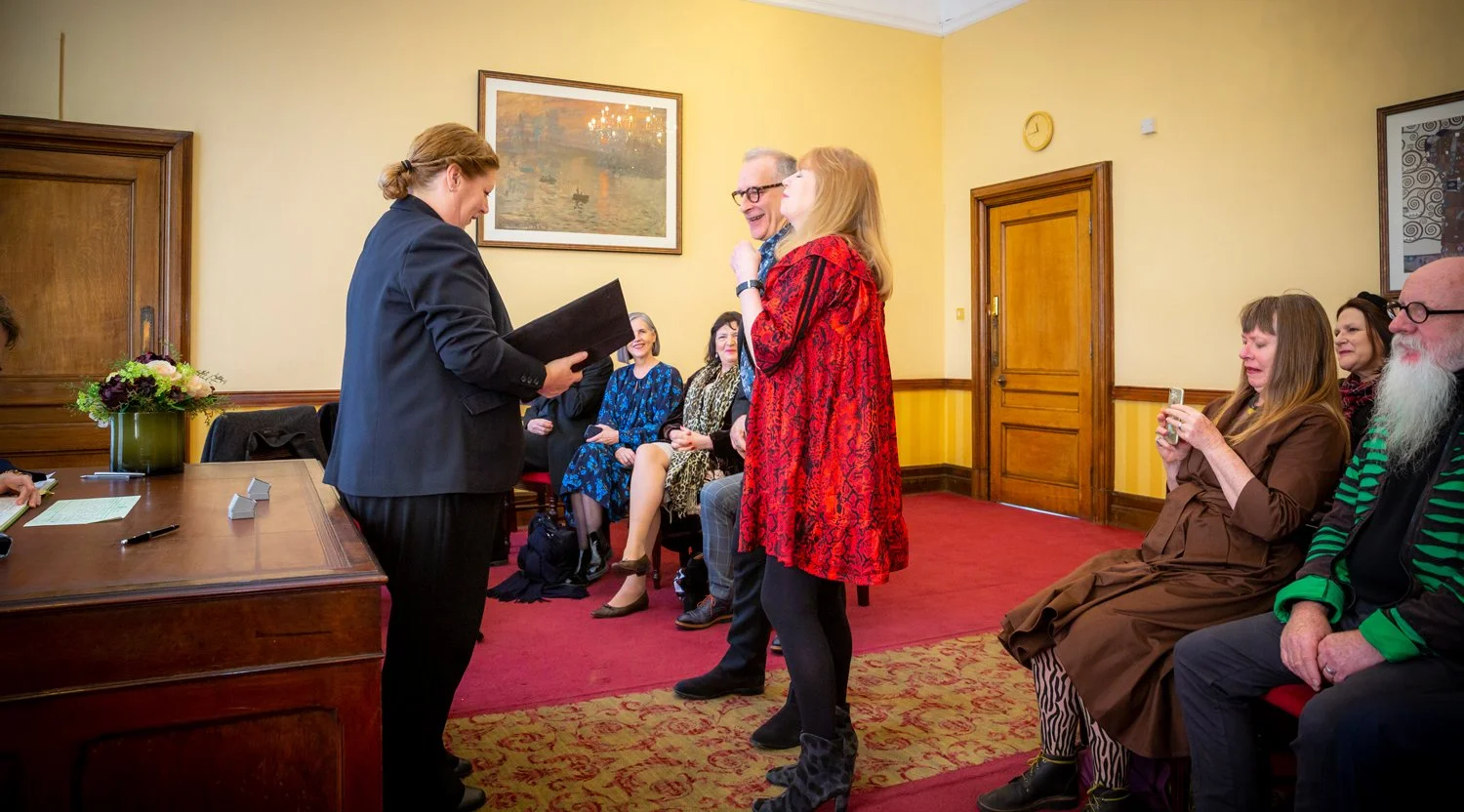 A woman in a blazer is reading from a folder to an older woman in a red dress, who is smiling and placing her hand on her chest. Several seated women and one seated man are watching, with some taking photos, in a room with yellow walls and framed art