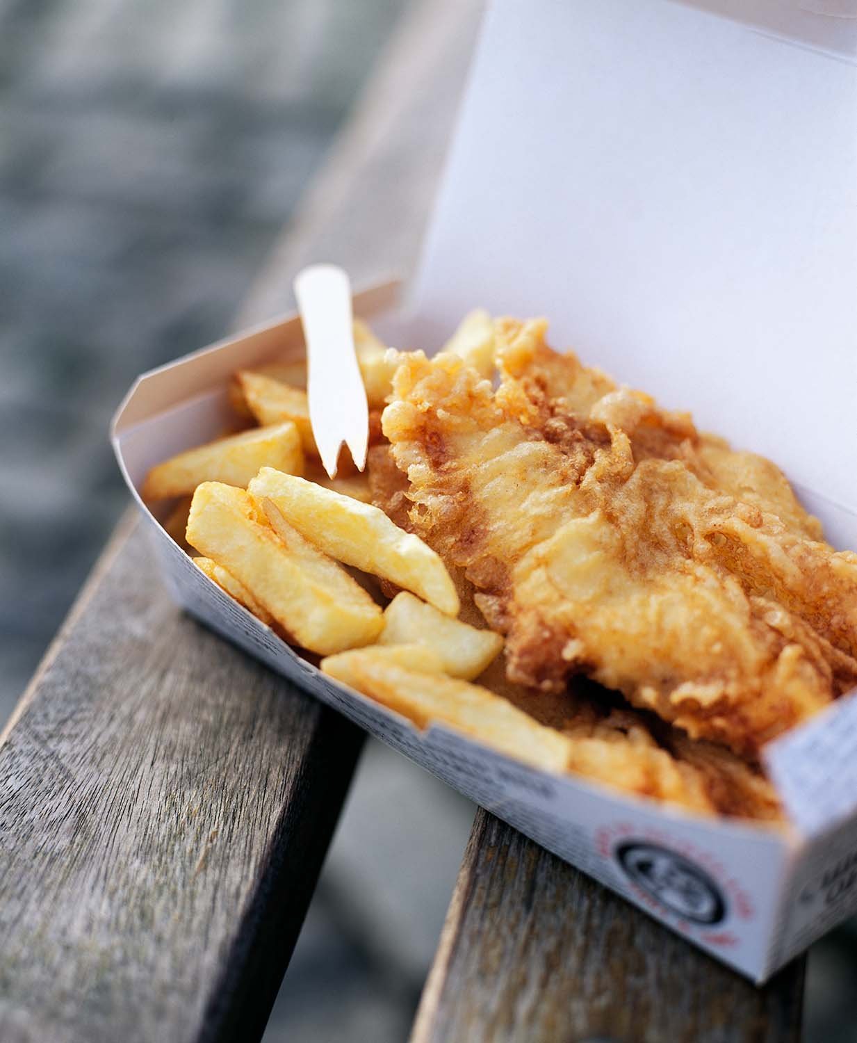 Close-up of a cardboard food container with fried fish and chips, placed on a wooden surface.