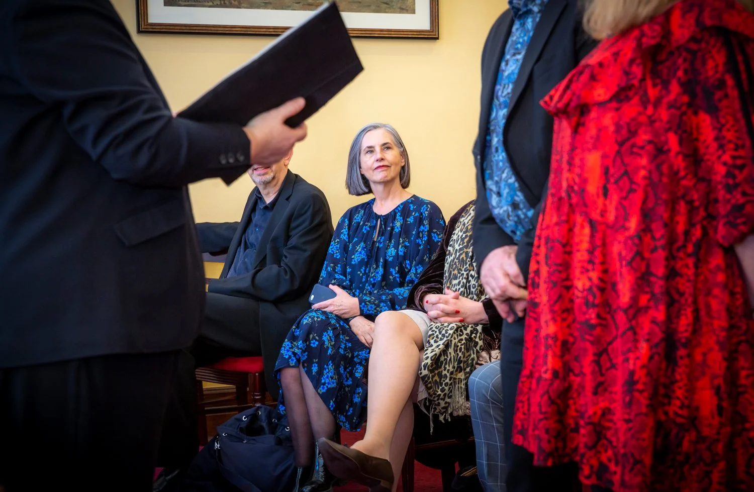 A group of people sitting and standing in a room with yellow walls, with a woman in a blue floral dress holding a phone, looking attentively at a person speaking or performing in front of her.