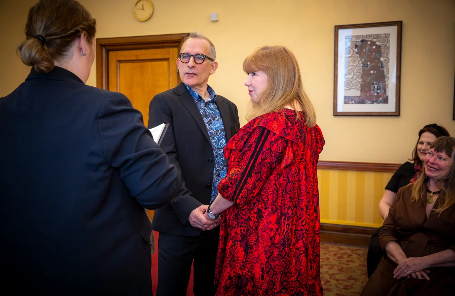 A woman in a red patterned dress holding hands with a man in a black suit and blue shirt, during a ceremony with women seated and watching in a room with yellow walls and framed art.