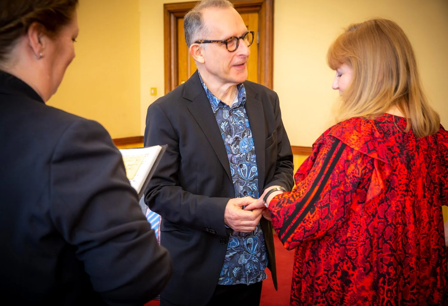 A man in a black suit and glasses holding hands with a woman in a red patterned dress during a formal event, with a woman in the background holding a notebook.