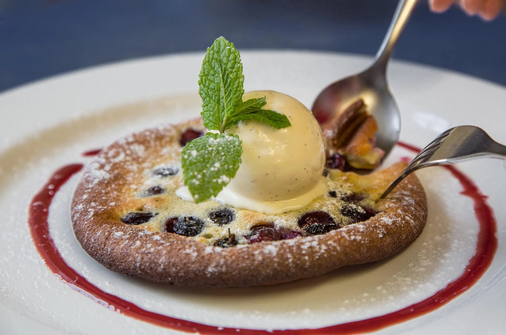 A dessert with a round pastry topped with a scoop of vanilla ice cream, garnished with mint leaves, powdered sugar, and a red fruit sauce, served on a white plate.