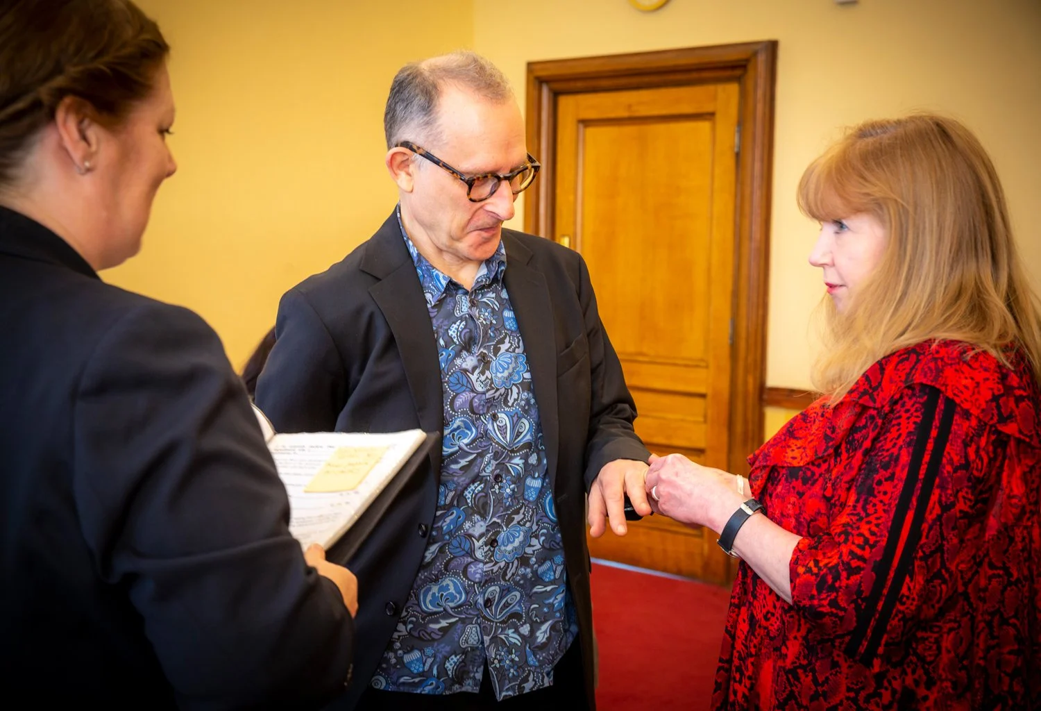 Three people, two women and one man, are engaged in a conversation indoors. The woman on the right with red hair is holding the man's hand. The second woman on the left, with dark hair, is holding a notepad.