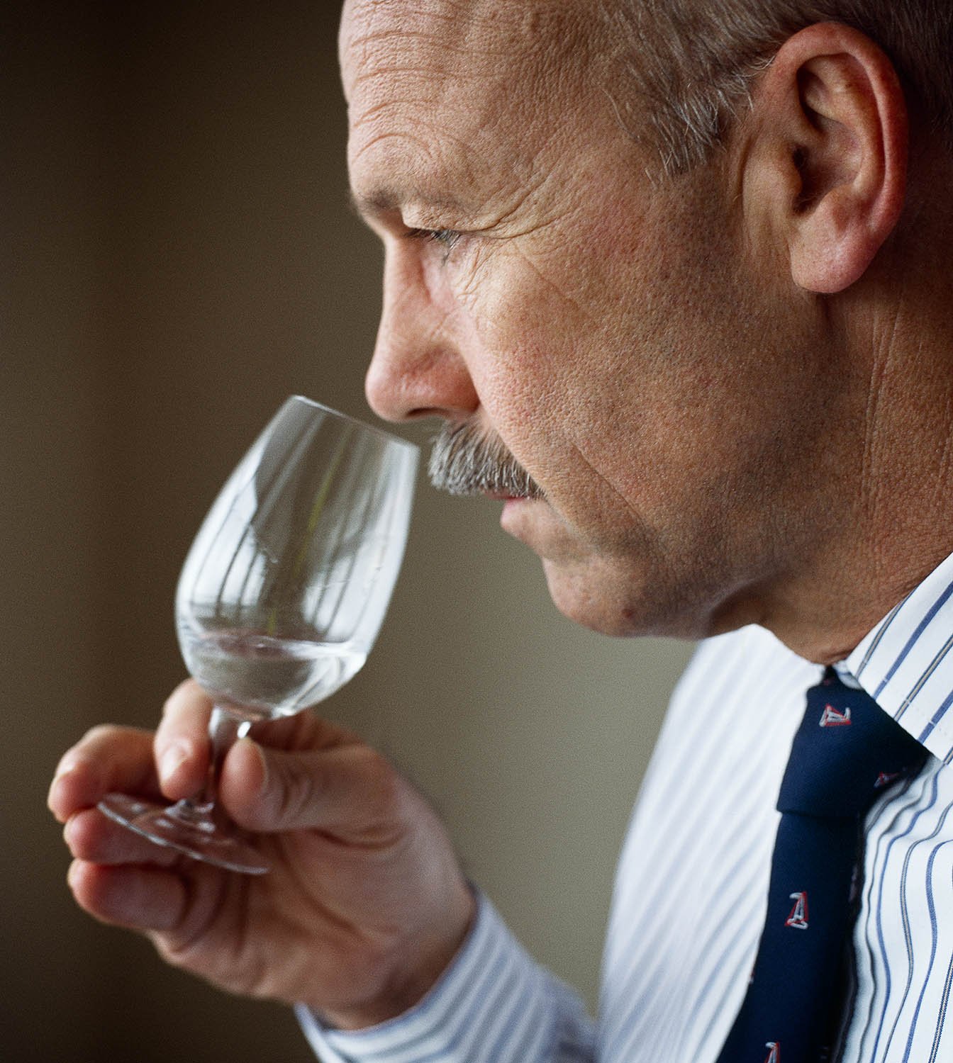 A middle-aged man with gray hair and a mustache smelling a glass of gin while holding it close to his nose.